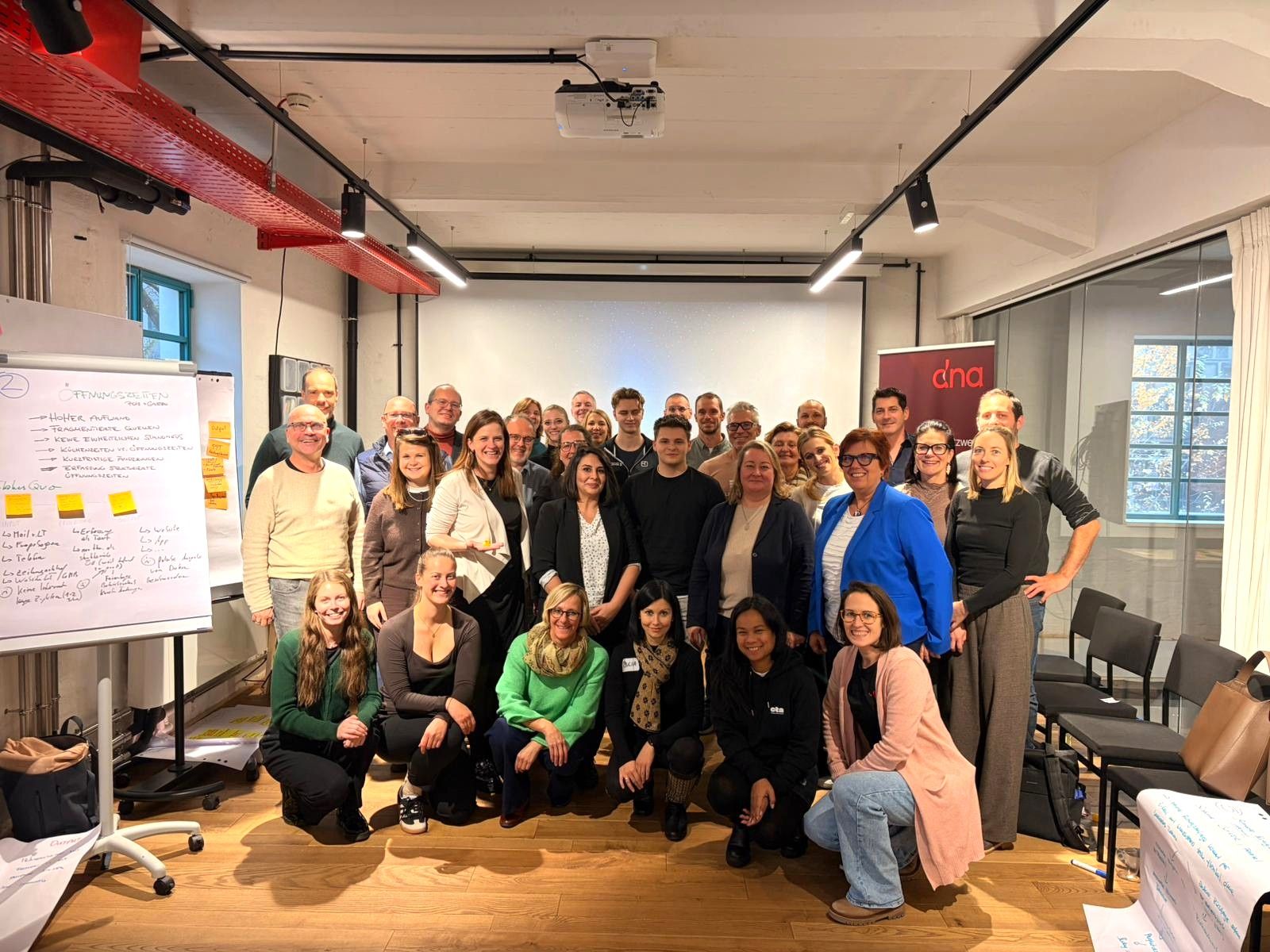 Groppenfoto eines Teams in einem Seminarraum: Rund 30 Personen stehen und knien für ein Foto; links ist ein Flipchart mit Notizzetteln zu sehen, im Hintergrund eine Projektionsfläche.