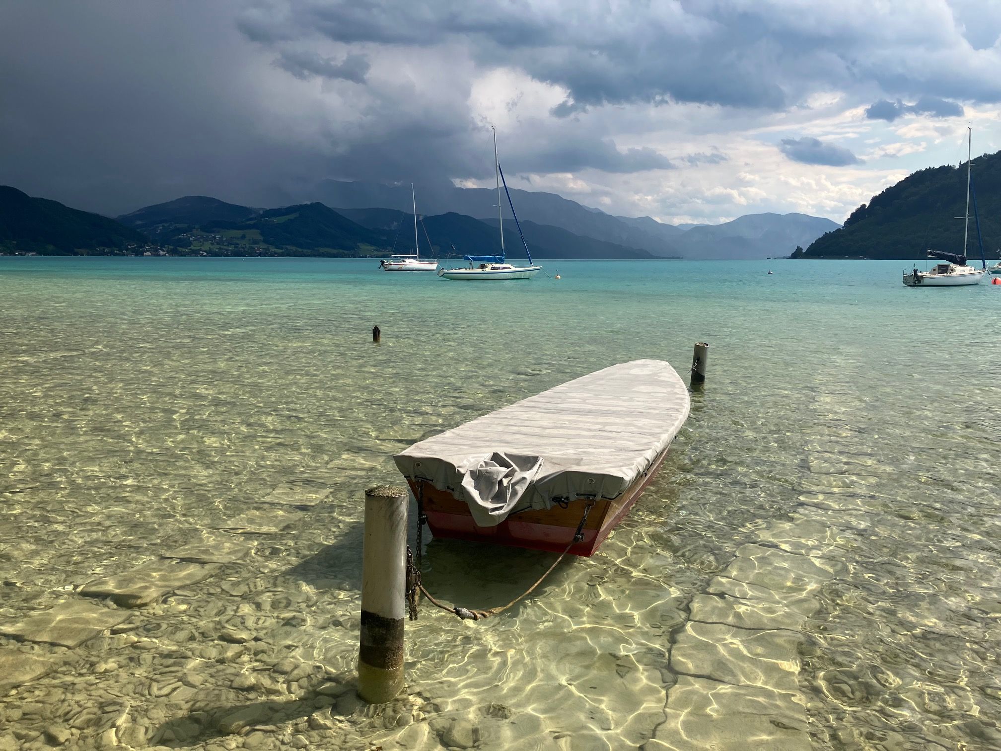 Ein kleines Boot liegt im klaren, seichten Wasser eines Sees; im Hintergrund sind Berge, Segelboote und ein bewölkter Himmel zu sehen.
