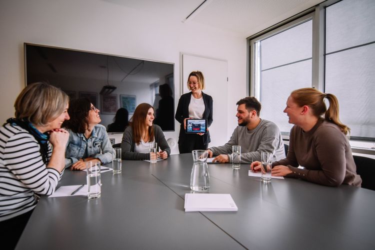 A group of colleagues is having a meeting, with one person holding a tablet displaying a video