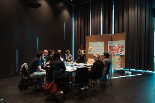 Gruppe von Personen arbeitet gemeinsam an einem Tisch in einem modernen Veranstaltungsraum mit Präsentationswand im Hintergrund.