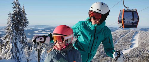 Zwei Skifahrerinnen in Winterkleidung mit Helmen und Skibrillen stehen auf einem verschneiten Berg; im Hintergrund sind schneebedeckte Bäume, eine weite Landschaft und eine Gondelbahn zu sehen.